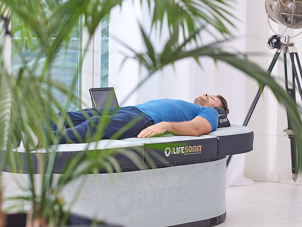 Das Bild zeigt eine Person, die auf einem medizinischen oder therapeutischen Bett liegt. Auf der Seite des Bettes ist der Markenname "LITECOM" sichtbar. Der Raum wirkt modern und gut beleuchtet, mit einigen grünen Pflanzen im Vordergrund und einer großen Lampe oder einem Scheinwerfer im Hintergrund.