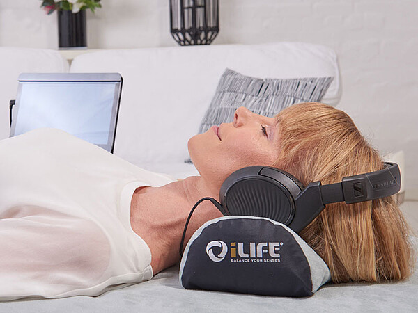 Das Bild zeigt eine Person, die auf einem Bett oder einer Couch liegt und große Over-Ear-Kopfhörer trägt. Unter dem Kopf der Person befindet sich ein schwarz-weißes Kissen oder Polster mit der Aufschrift "iLIFE" und "Digital Therapy System". Im Hintergrund steht ein offener Laptop auf einer Oberfläche hinter der Person.